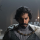 Dev Patel's Gawain in The Green Knight has his head bowed and is wearing several layers of grey with a gold chain holding his cloak. Two armor clad knights stand behind him on either side.